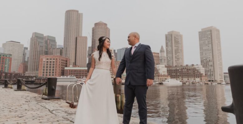 A couple holding hands, stands by a waterfront with Boston’s skyline in the background