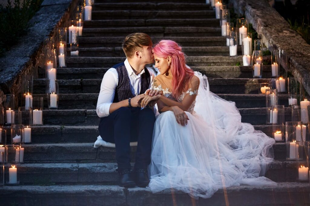 Bride with pink hair and stylish groom sit on footsteps with shiny candles