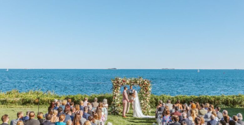 A wedding ceremony by the sea, guests seated, vows exchanged under a floral arch