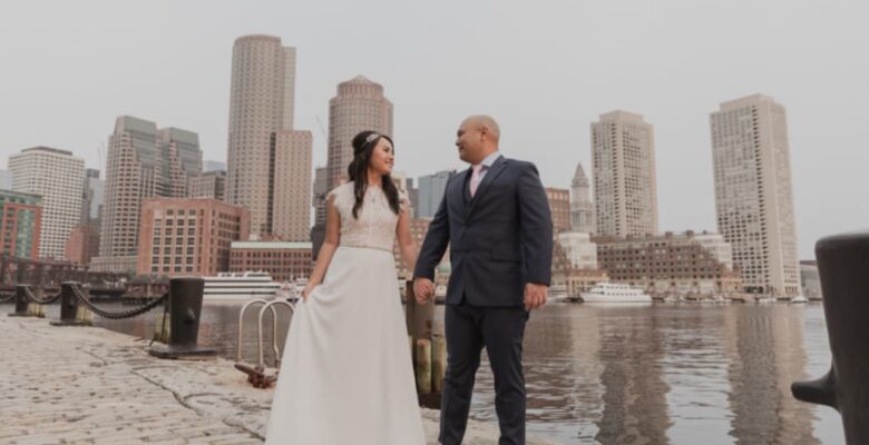 A couple holding hands, stands by a waterfront with Boston’s skyline in the background